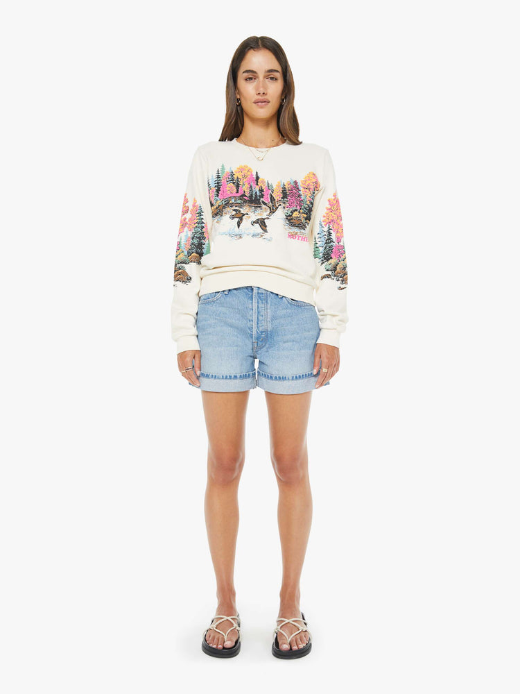 Full body close up view of a woman in an off white sweatshirt features an idyllic lakeside graphic with MOTHER's name in pink on the front.