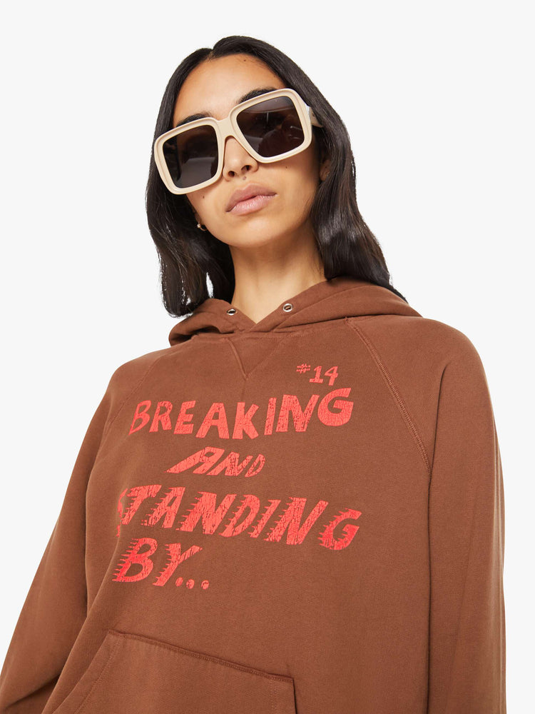 Close up view of a woman in brown with a faded text graphic hoodie with an oversized patch pocket and a loose, comfortable fit.
