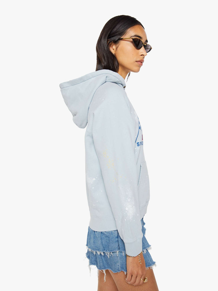 Side view of a woman in a light blue, the hoodie features a crackle-printed scrap yard graphic on the front with paint and bleach splatters throughout.