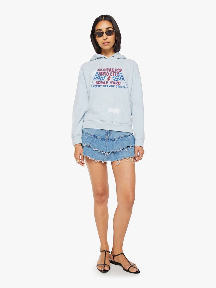 Full body view of a woman in a light blue, the hoodie features a crackle-printed scrap yard graphic on the front with paint and bleach splatters throughout.