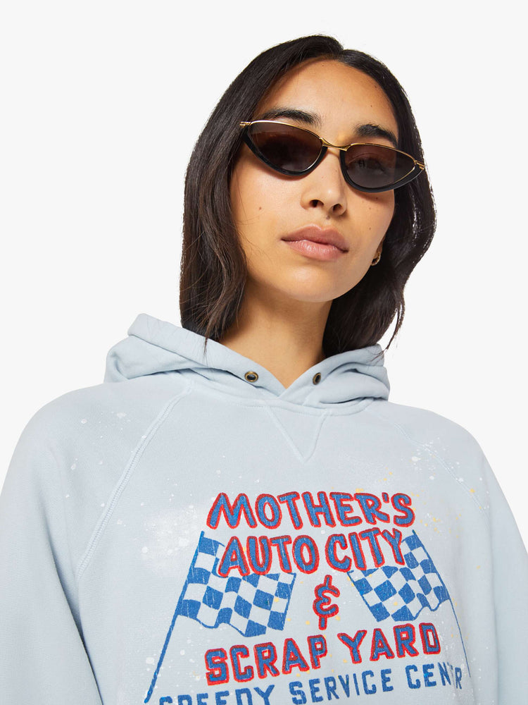 Close up view of a woman in a light blue, the hoodie features a crackle-printed scrap yard graphic on the front with paint and bleach splatters throughout.