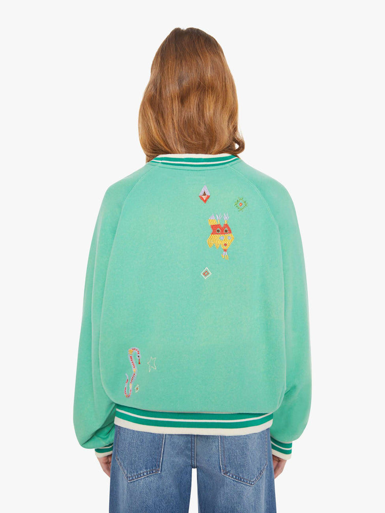 Back view of a woman in a sun-faded green sweatshirt with colorful embroidery throughout and the number 15 appliqued on the front.