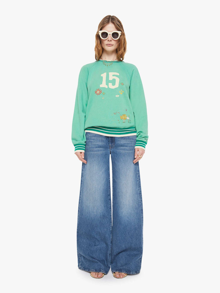 Full body view of a woman in a sun-faded green sweatshirt with colorful embroidery throughout and the number 15 appliqued on the front.