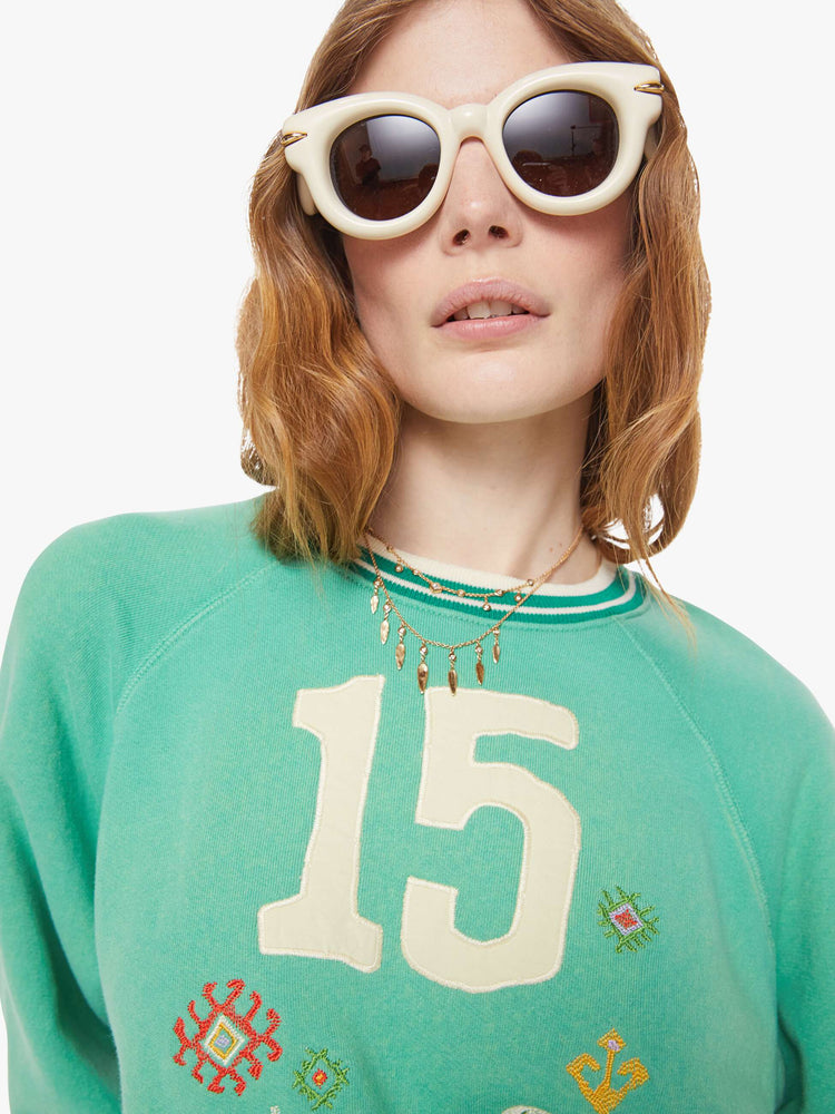 Swatch view of a woman in a sun-faded green sweatshirt with colorful embroidery throughout and the number 15 appliqued on the front.