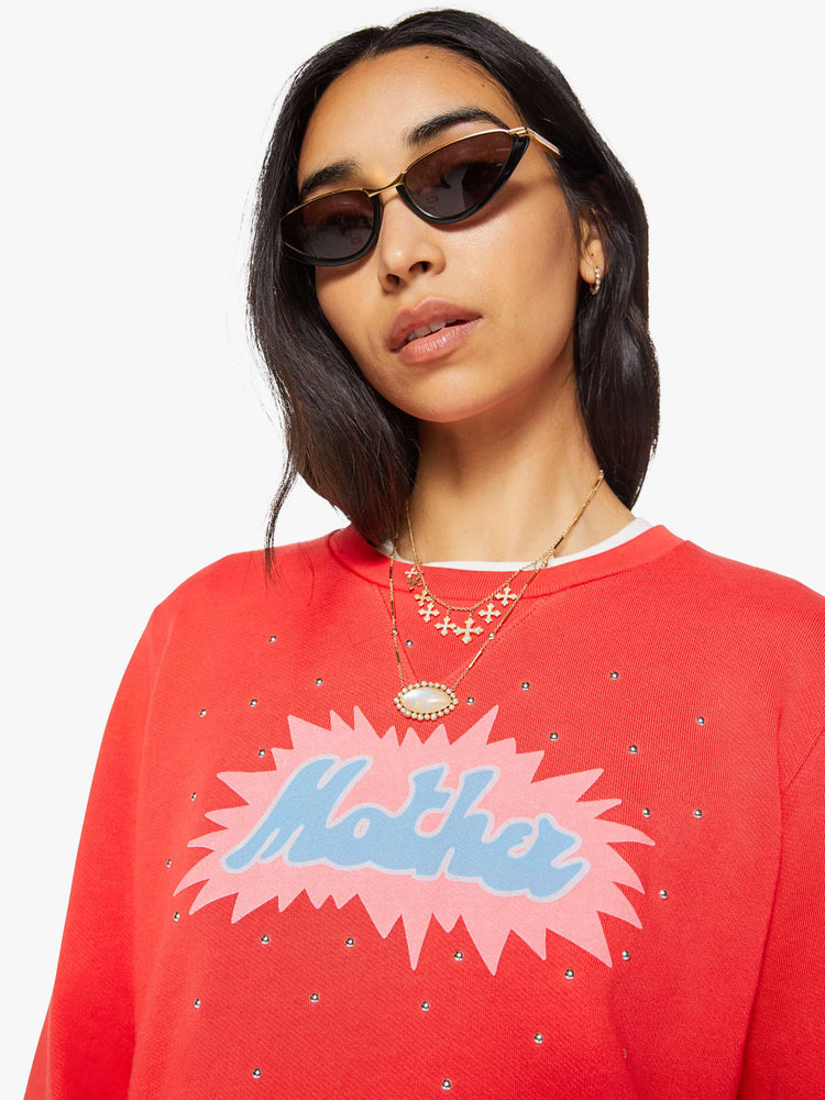 Close up view of a woman in a sweatshirt is designed in faded red with a pink and light blue printed graphic and small metal studs throughout.