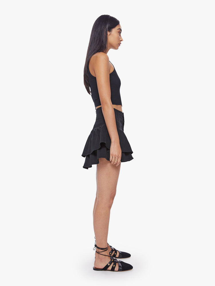 Side view of a woman denim mini skirt with a high rise, exposed button fly, snug waist and ruffles along the hem and diagonally across the front in a solid black hue.