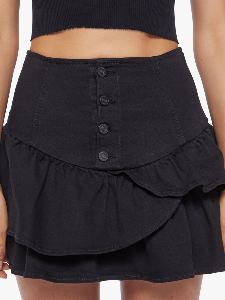 Close up view of a woman denim mini skirt with a high rise, exposed button fly, snug waist and ruffles along the hem and diagonally across the front in a solid black hue.