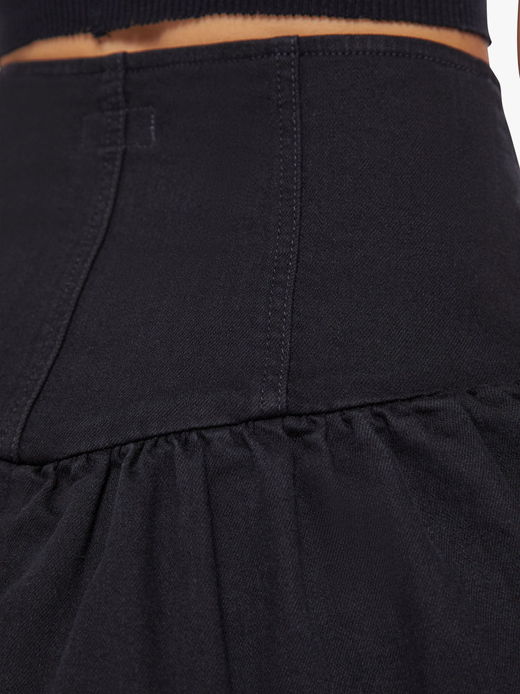 Back waist close up view of a woman denim mini skirt with a high rise, exposed button fly, snug waist and ruffles along the hem and diagonally across the front in a solid black hue.