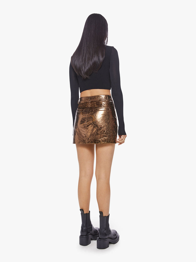 Back view of a woman high shine metallic golden hue zip-up mini skirt with a high rise, buttoned waistband and patch pockets.