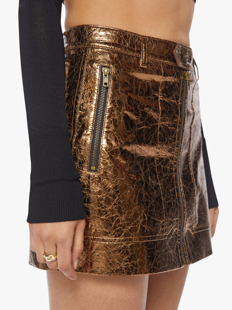 Close up view of a woman high shine metallic golden hue zip-up mini skirt with a high rise, buttoned waistband and patch pockets.