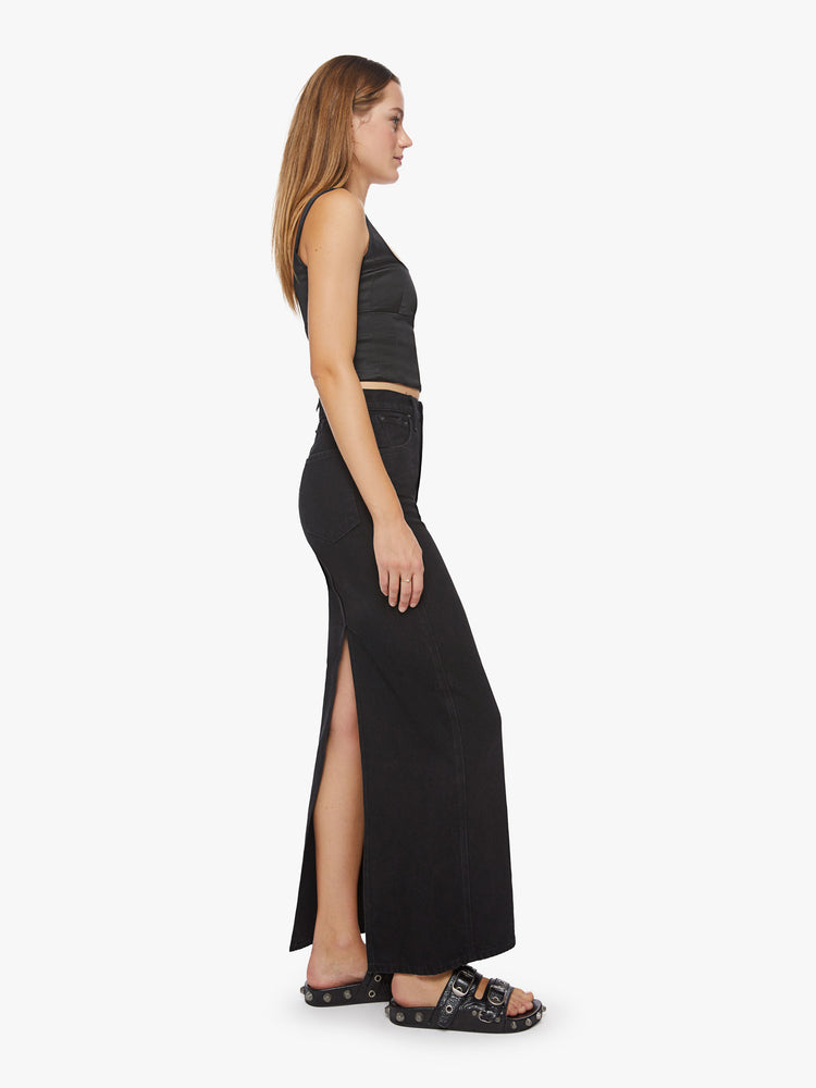 Side view of a woman black denim skirt with a high rise, floor-length hem and a back slit.