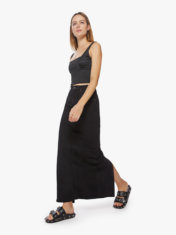 Walking view of a woman black denim skirt with a high rise, floor-length hem and a back slit.