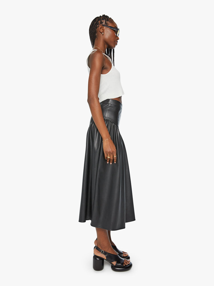 Side view of a woman midi skirt fitted at the waist with diagonal seams and ruffles for a flowy fit in a black faux leather.