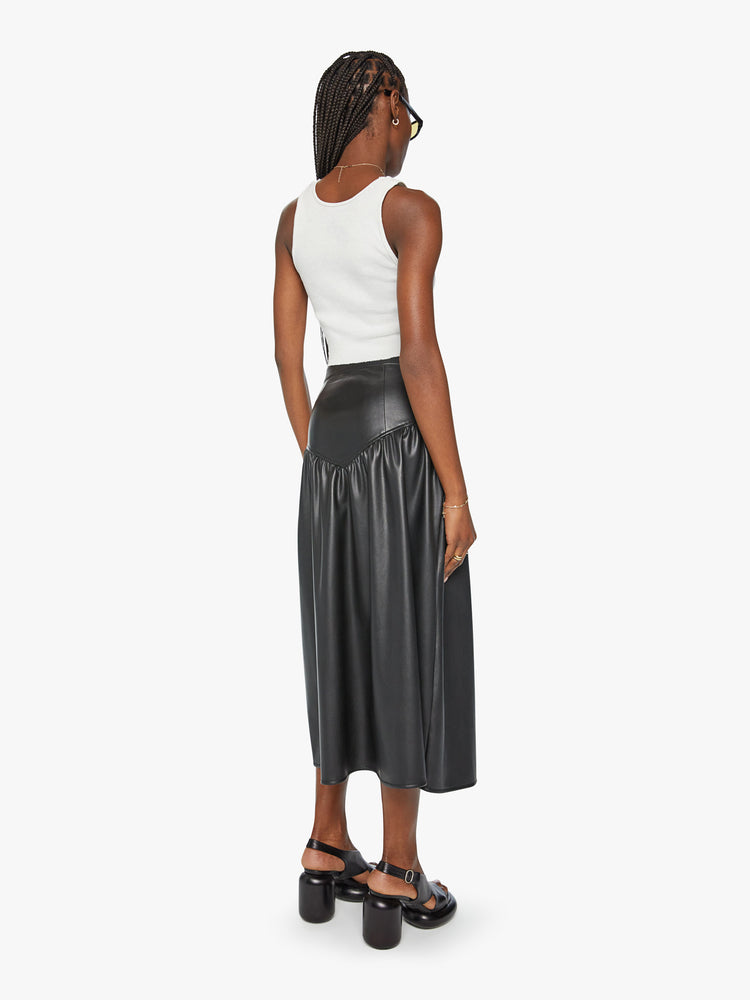 Back view of a woman midi skirt fitted at the waist with diagonal seams and ruffles for a flowy fit in a black faux leather.