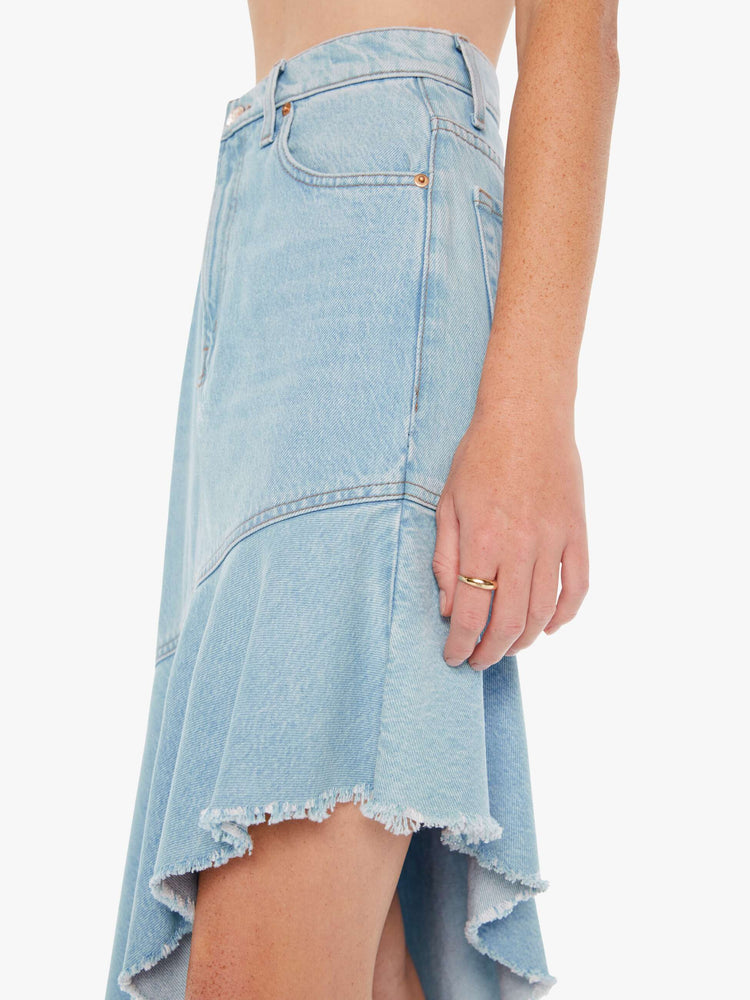 Side close up view of a woman wearing a light blue wash skirt featuring a super high rise and and exaggerated asymmetrical ruffled raw hem.