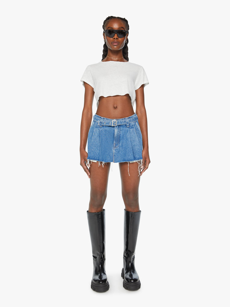 Front view of a woman denim mini skirt with a belted waistband, wide front pleats and a super-short frayed hem with exposed pocket lining in a mid blue wash.