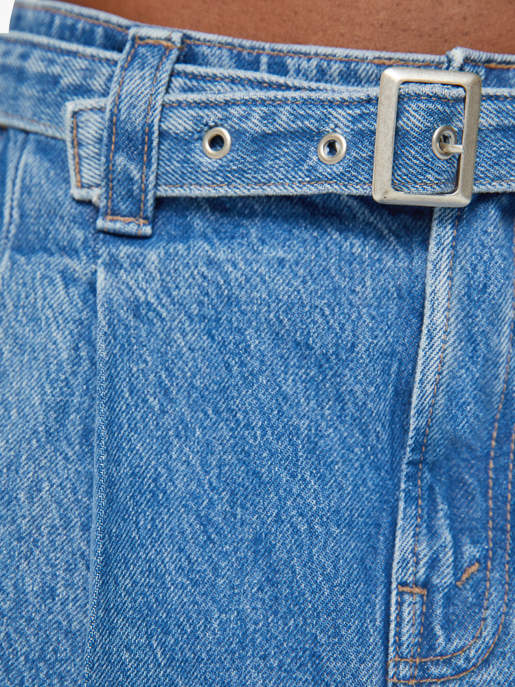 Swatch view of a woman denim mini skirt with a belted waistband, wide front pleats and a super-short frayed hem with exposed pocket lining in a mid blue wash.