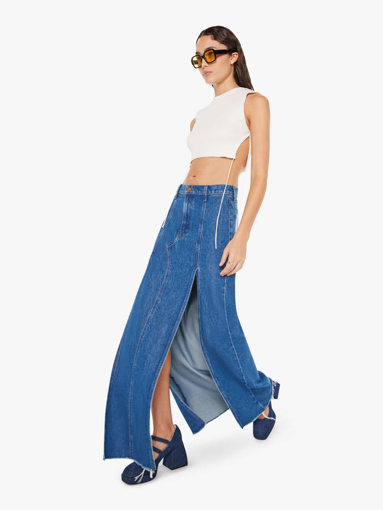 Walking view of a woman mid blue wash high-rise skirt is designed with thigh-high slits up the front, a frayed hem and a flowy fit.