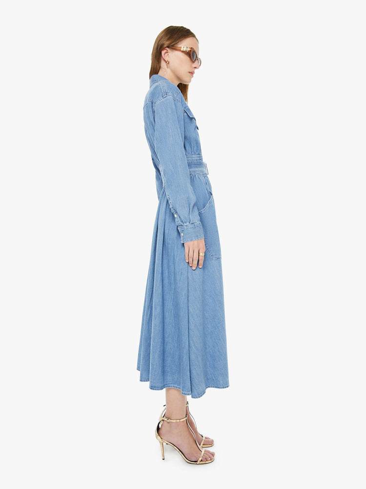 Side view of a woman in a  denim prairie maxi dress with a collared neck, puffed long sleeves, a fitted waist with a detachable belt, patch pockets and faux-pearl snaps down the front. 