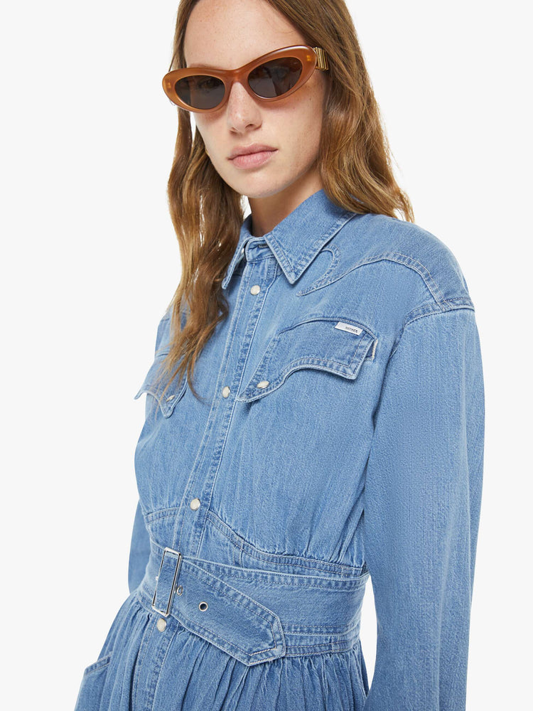 Close up view of a woman in a  denim prairie maxi dress with a collared neck, puffed long sleeves, a fitted waist with a detachable belt, patch pockets and faux-pearl snaps down the front. 