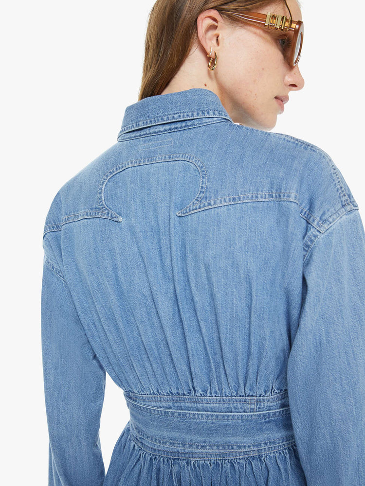 Back view of a woman in a  denim prairie maxi dress with a collared neck, puffed long sleeves, a fitted waist with a detachable belt, patch pockets and faux-pearl snaps down the front. 