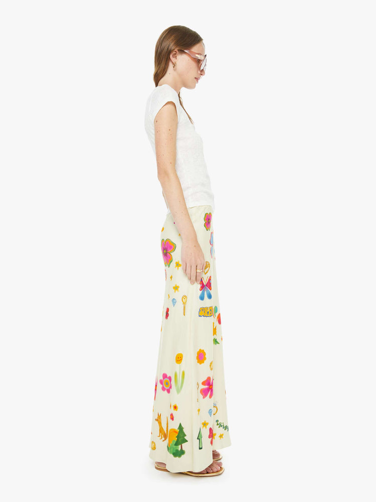 Side view of a woman in maxi slip skirt with a mid rise, loose fit and an ankle-length hem in offwhite with colorful flowers, stars, butterflies, leaves and more throughout.