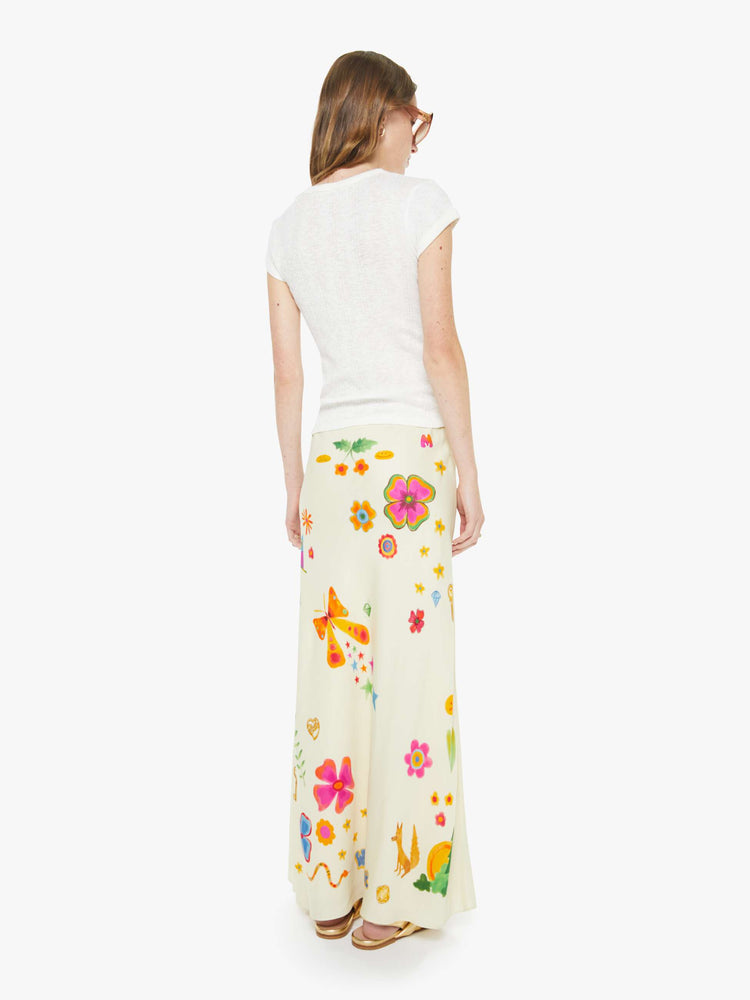 Back view of a woman in maxi slip skirt with a mid rise, loose fit and an ankle-length hem in offwhite with colorful flowers, stars, butterflies, leaves and more throughout.