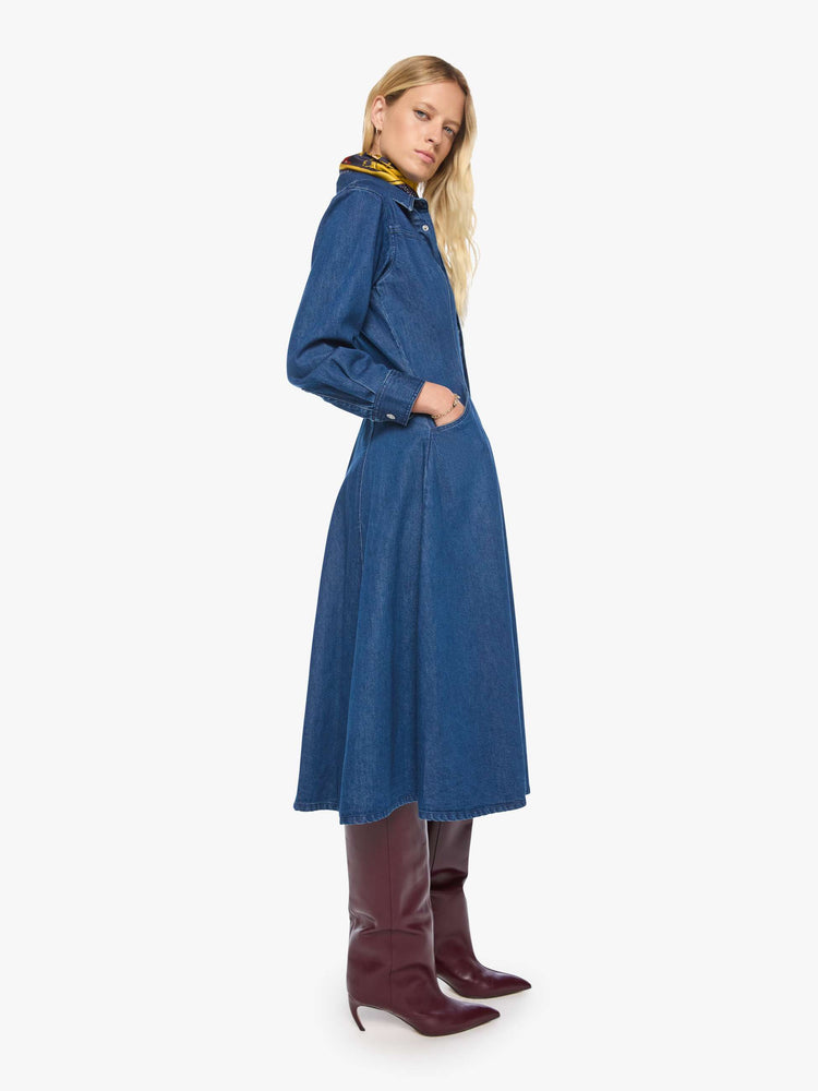 Side view of a woman in a dark blue denim shirtdress with a pointed yoke, long balloon sleeves, slit pockets, buttons down the front and a flared midi skirt.