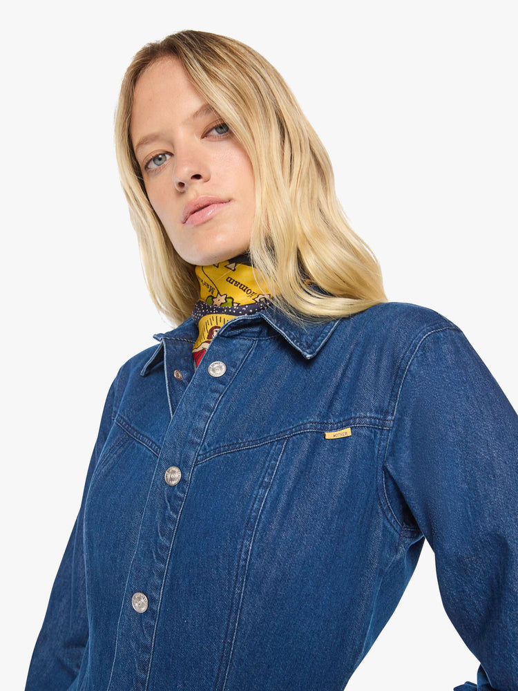 Close up view of a woman in a dark blue denim shirtdress with a pointed yoke, long balloon sleeves, slit pockets, buttons down the front and a flared midi skirt.
