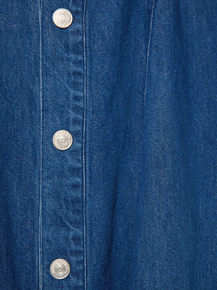 Button view of a woman in a dark blue denim shirtdress with a pointed yoke, long balloon sleeves, slit pockets, buttons down the front and a flared midi skirt.