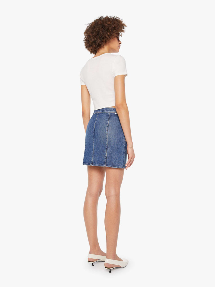 Back view of a woman in mid-blue high-waisted denim skirt with a zip fly, patch pockets with rivet details, a seam down the front and thigh-length hem.