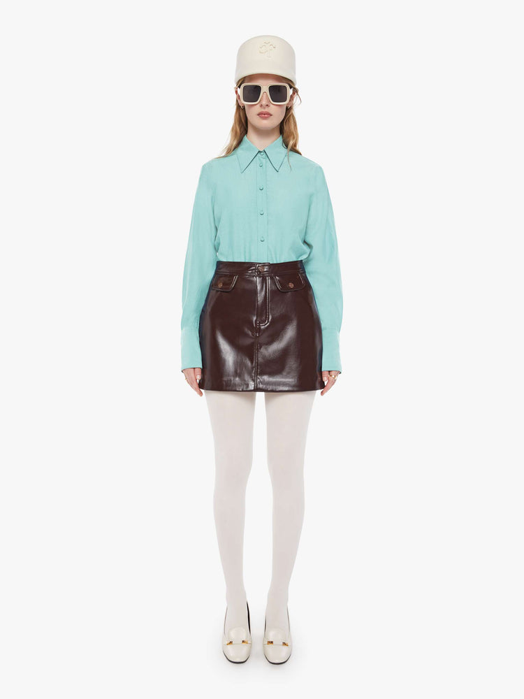 Front view of a woman in a chocolate brown faux patent leather mini skirt with a high rise, button fly, small pockets at the waist and a short hem. 