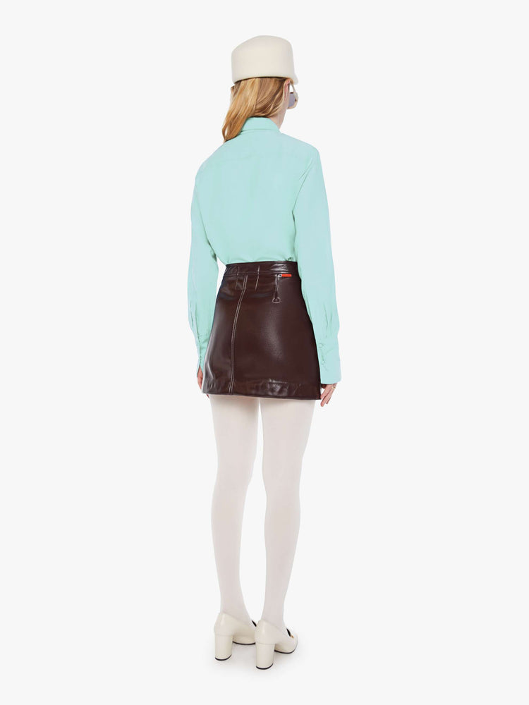 Back view of a woman in a chocolate brown faux patent leather mini skirt with a high rise, button fly, small pockets at the waist and a short hem. 