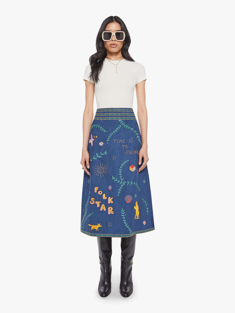 Front view of a woman in an A-line skirt with a high rise, side-zip closure and a calf-length hem in a deep navy with colorful embroidered leaves, text and doodles throughout.