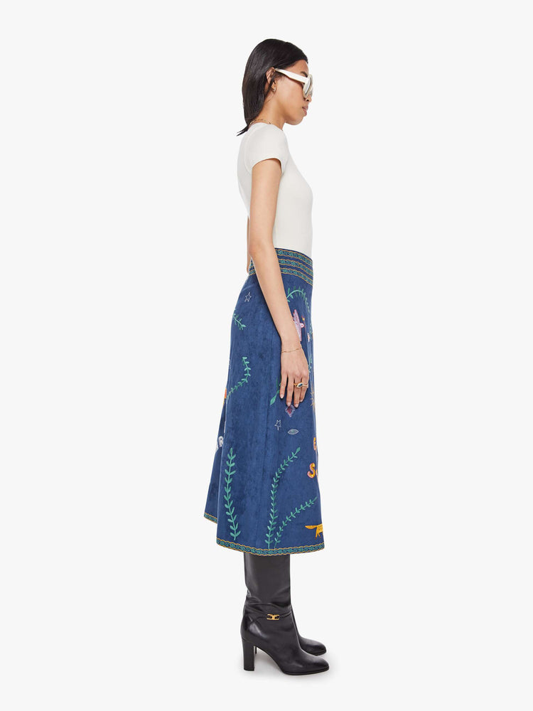 Side view of a woman in an A-line skirt with a high rise, side-zip closure and a calf-length hem in a deep navy with colorful embroidered leaves, text and doodles throughout.