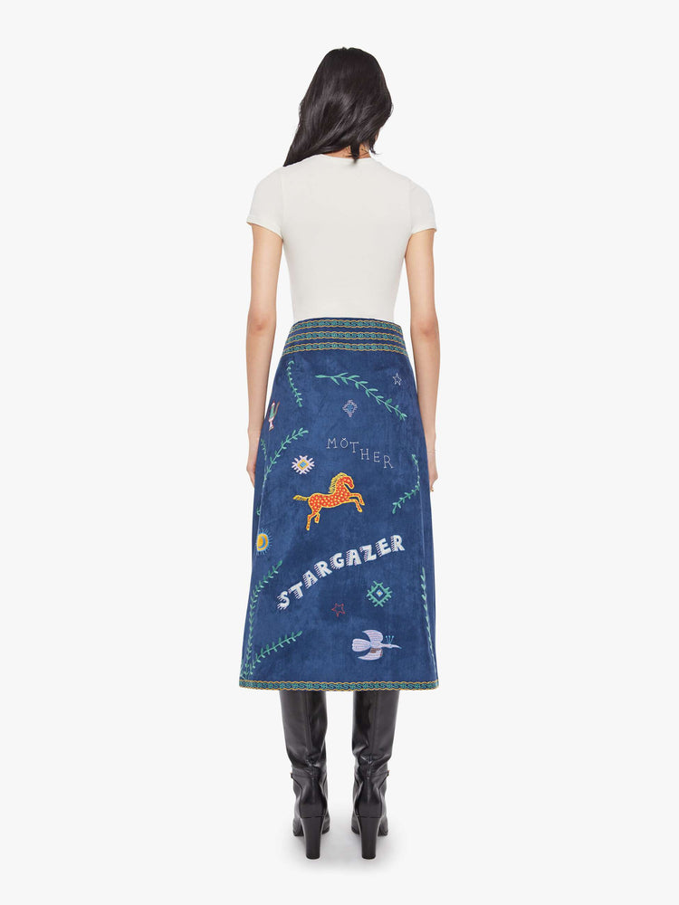 Back view of a woman in an A-line skirt with a high rise, side-zip closure and a calf-length hem in a deep navy with colorful embroidered leaves, text and doodles throughout.