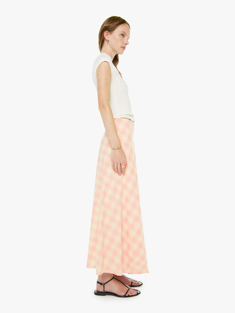 Side view of a woman in a cream and baby pink gingham print flowy maxi skirt with a tied waistband, ankle-length hem and a flowy A-line fit.