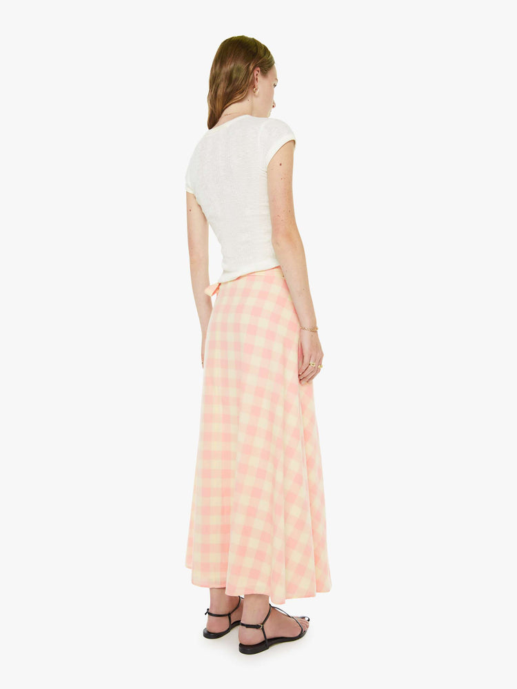 Back view of a woman in a cream and baby pink gingham print flowy maxi skirt with a tied waistband, ankle-length hem and a flowy A-line fit.