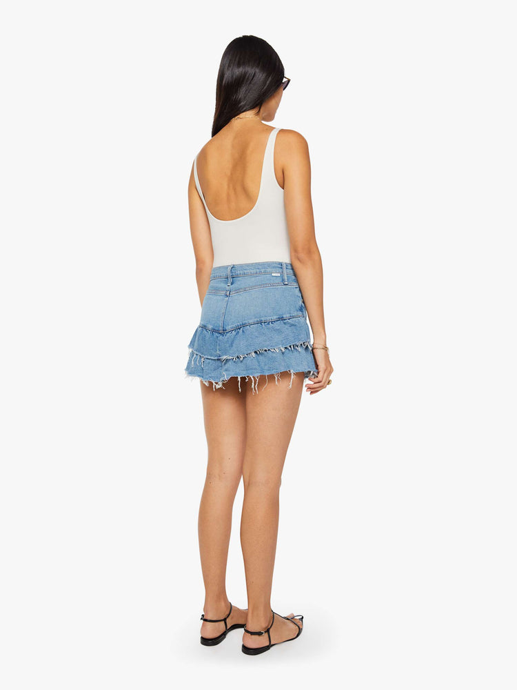 Back view of a woman in a light blue denim mini skirt with a zip fly, angled ruffles and a frayed hem.
