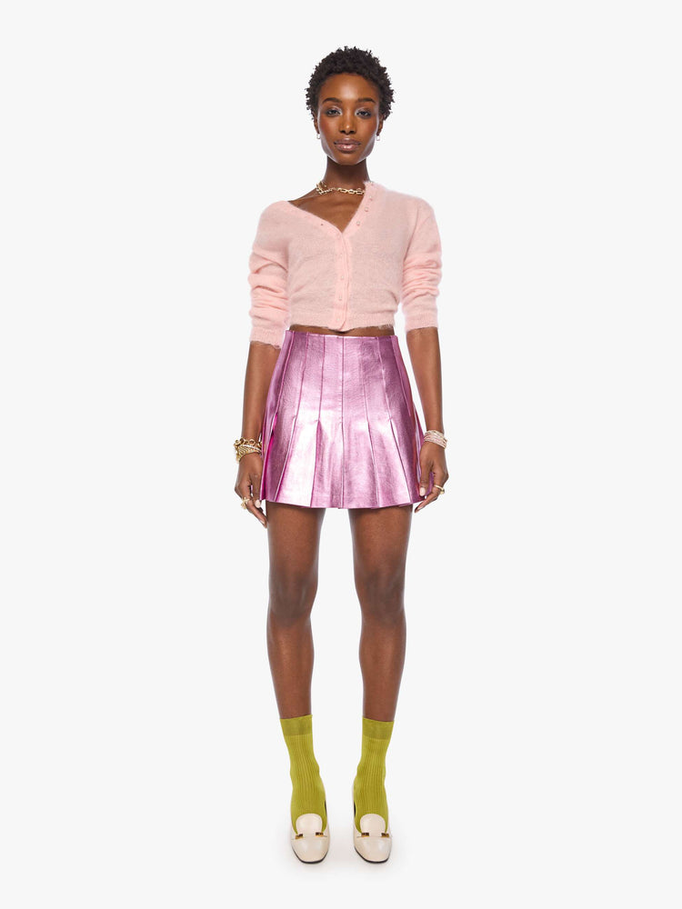Front view of a woman in a reflective light pink faux leather pleated mini skirt with a high rise, wide pleats and a thigh-grazing hem. 