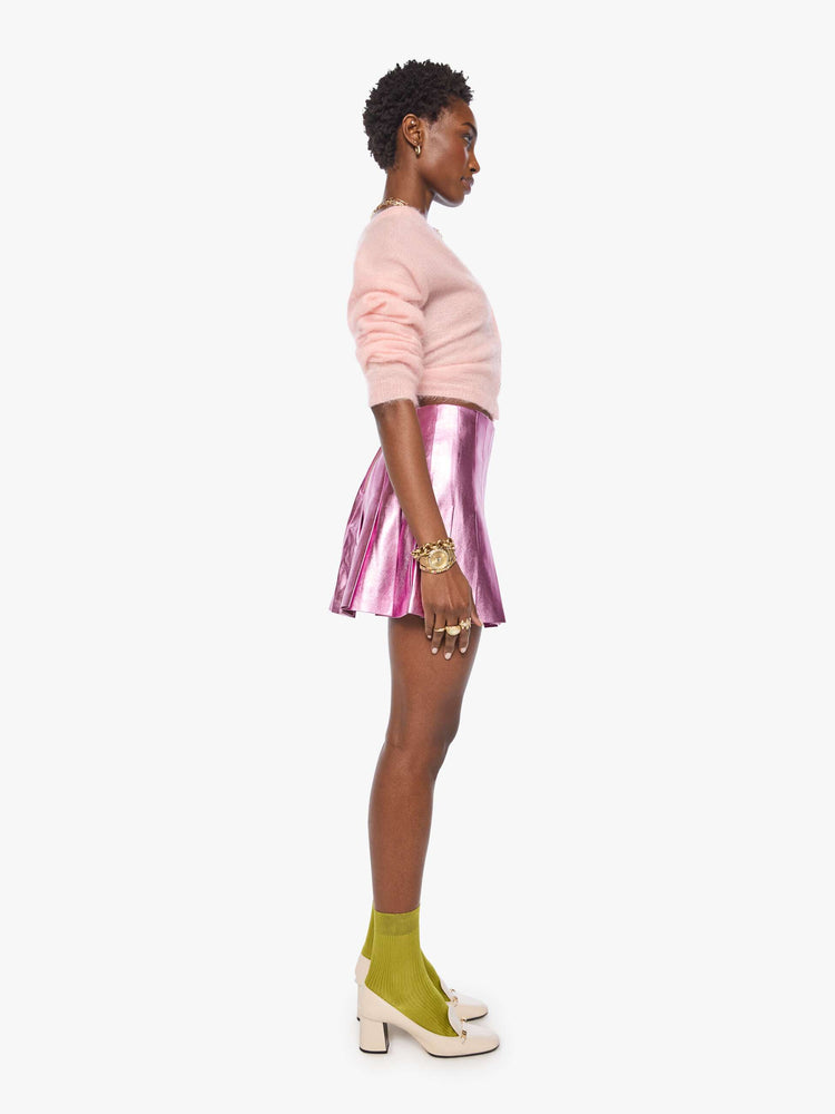 Side view of a woman in a reflective light pink faux leather pleated mini skirt with a high rise, wide pleats and a thigh-grazing hem. 