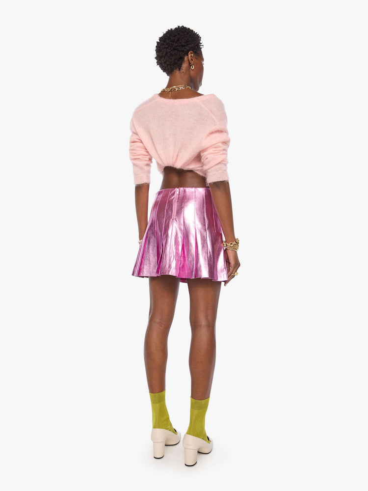 Back view of a woman in a reflective light pink faux leather pleated mini skirt with a high rise, wide pleats and a thigh-grazing hem. 