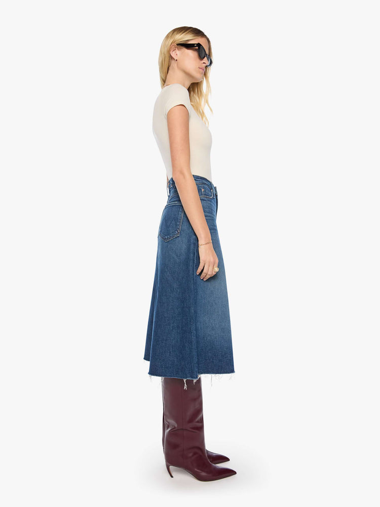 Side view of a woman in a mid blue denim midi skirt with a zip fly, frayed hem and an A-line fit.