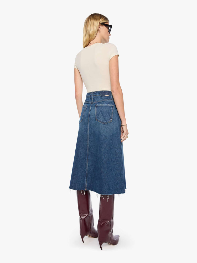Back view of a woman in a mid blue denim midi skirt with a zip fly, frayed hem and an A-line fit.