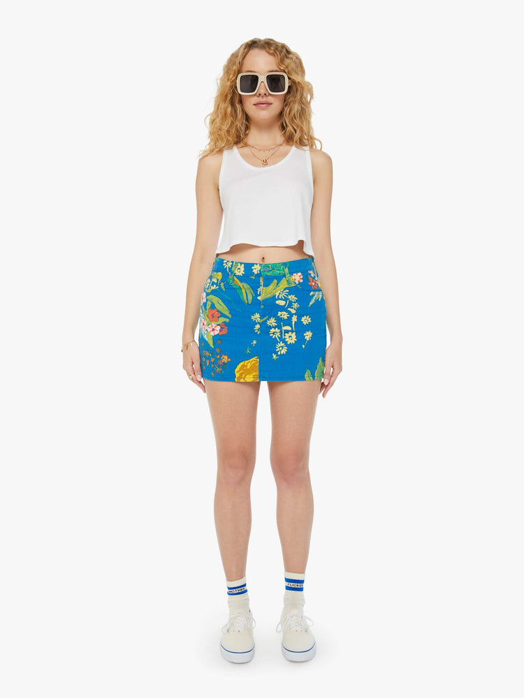 Front view of a woman in a bright blue mini skirt with an oversized tropical floral print, high rise, button fly, slim fit and thigh-grazing hem. 
