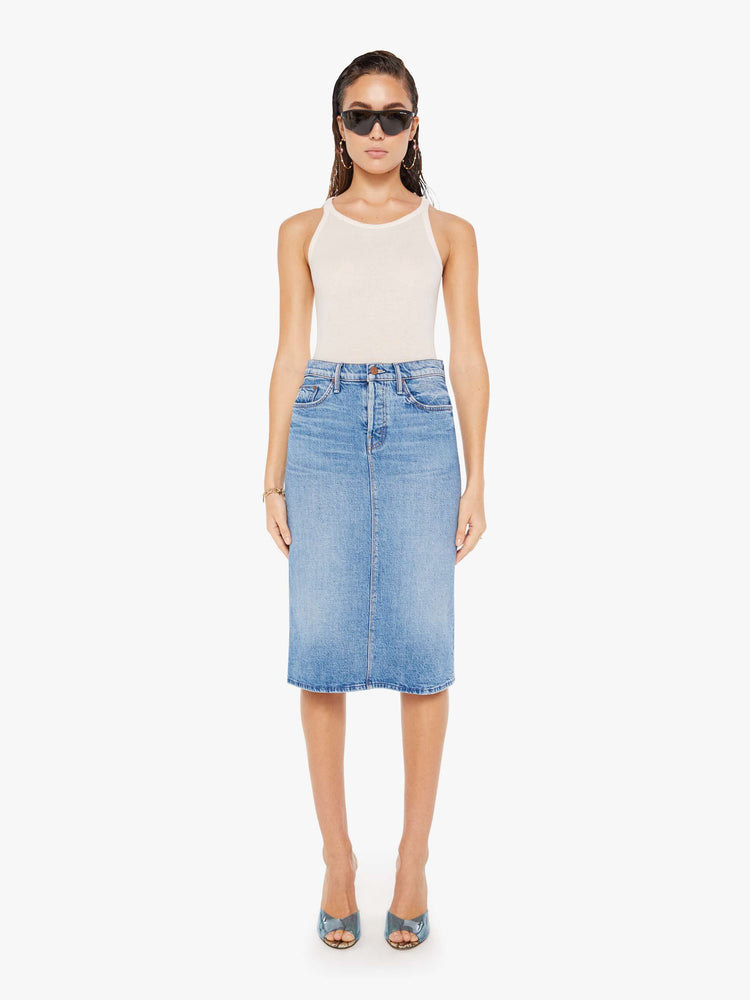 Front view of a woman wearing a medium blue wash denim midi skirt featuring a relaxed mid rise, paired with a white tank top.