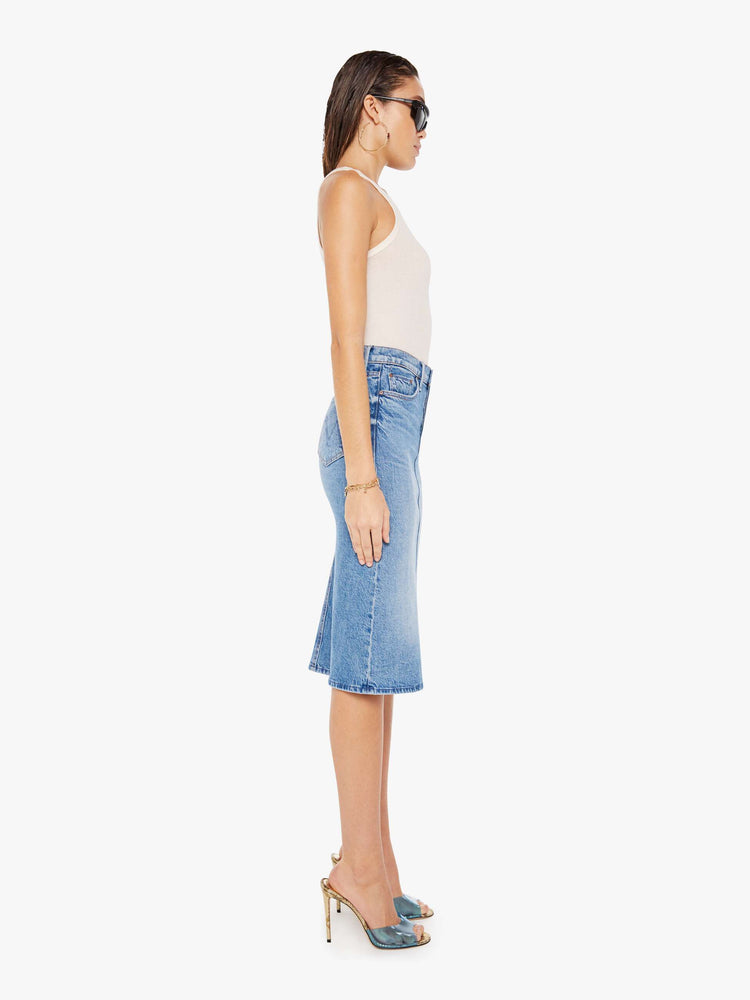 Side view of a woman wearing a medium blue wash denim midi skirt featuring a relaxed mid rise, paired with a white tank top.