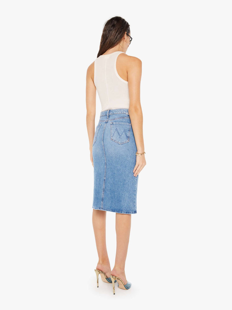 Back view of a woman wearing a medium blue wash denim midi skirt featuring a relaxed mid rise, paired with a white tank top.