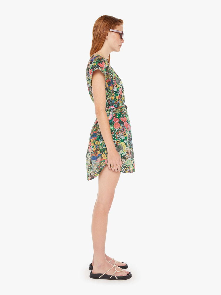 A side full body view of a woman wearing a short sleeve shirt dress, featuring a colorful garden print and a waist tie.