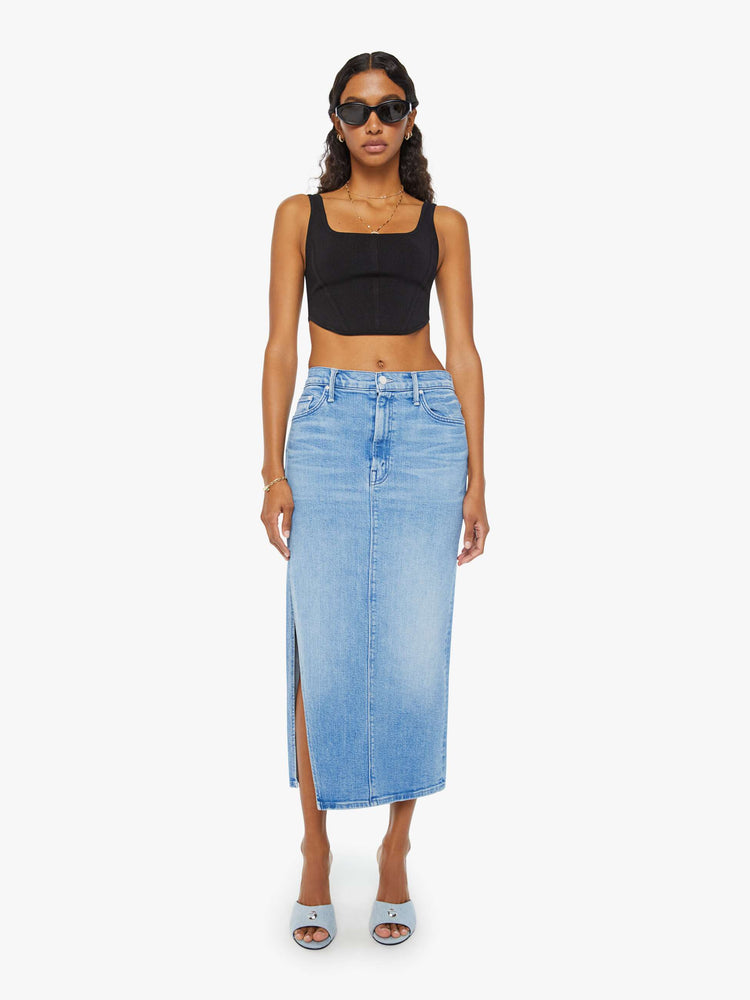 Front view of a woman in a light blue high-rise denim midi skirt with a diagonal thigh-high slit and a narrow fit with whiskering and fading at the knees. Styled with a black tank top.